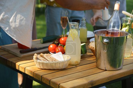 Plan rapproché d'une table d'appoint par grill lors d'une garden party estivale. Table de travail avec vin, légumes frais, pain et couteau.