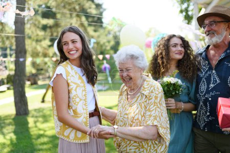 Photo for Beautiful senior birthday woman receiving gift from granddaughter. Two mature sisters with grandparents at birthday party. Multigenerational family. - Royalty Free Image