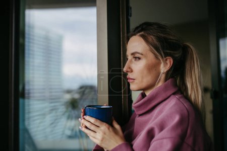 Foto de Mujer disfrutando de una taza de té caliente, café en casa después del trabajo, de pie junto a la ventana con capucha rosa. Equilibrio entre vida laboral y personal sanitario. Perforaciones nasales. - Imagen libre de derechos