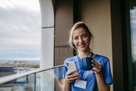 Photo for Nurse, doctor in uniform taking break, standing on hospital balcony and drinking coffee, scrolling on smartphone. Staff break area. Work-life balance of healthcare worker. - Royalty Free Image