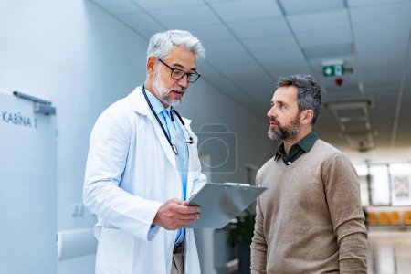 Doctor hablando con el paciente en el pasillo del hospital. Guapo médico con cabello gris sujetando portapapeles con resultado de la prueba, consultando con el hombre, de pie en la clínica privada moderna.