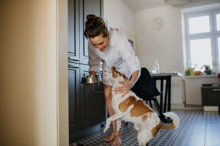Téléchargez les photos : Femme en pyjama nourrissant le chien, souriant. La routine du week-end à la maison. Hygge style de vie pour femme célibataire. - en image libre de droit