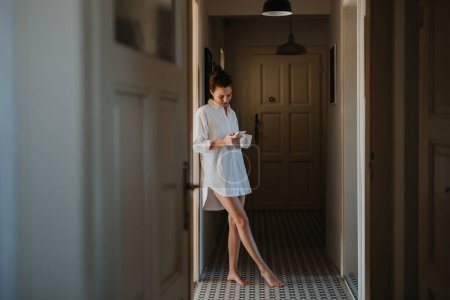 Foto de Mujer joven en pijama por la mañana, llevando una taza de café y volviendo a la cama. Vista lateral de la hermosa mujer soltera en casa sola durante el fin de semana. - Imagen libre de derechos