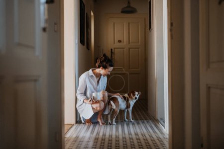 Foto de Mujer en pijama, jugando con su lindo perro, bebiendo una taza de café de la mañana. Relajación de fin de semana en casa. Estilo de vida Hygge. - Imagen libre de derechos