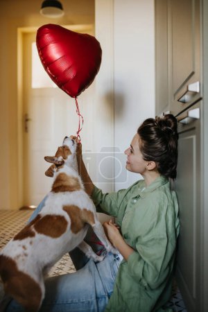 Beautiful single woman sitting on floor, playing with her dog with balloon. Young woman living alone in apartment, enjoying weekend.