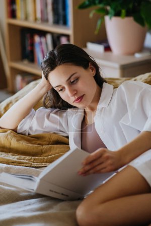 Foto de Hermosa mujer leyendo libro en la cama, sensación acogedora. Libertad, actividad de fin de semana para mujer soltera. - Imagen libre de derechos