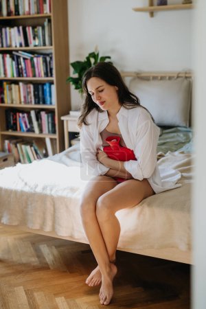 Junge Frau zu Hause, die unter Menstruationsschmerzen leidet und Krämpfe hat. Frau wärmt Unterbauch mit Wärmflasche, Endometriose und Schmerzen im Bauch.