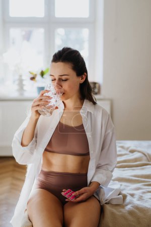 Photo for Woman taking pills with water, pills, suffering from menstrual pain, having cramps. Close up of woman holding abdomen, endometriosis, and conditions causing pain in tummy. - Royalty Free Image