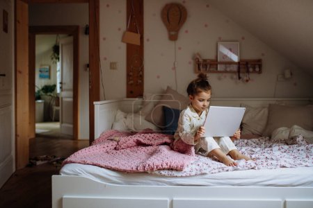 Foto de Linda chica sentada en la cama y viendo algo en la tableta. Tiempo de pantalla de los niños. - Imagen libre de derechos