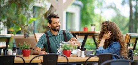Photo for Young couple at date in restaurant, sitting on restaurant terrace. Boyfriend pouring water in girlfriends glass, taking care of her. Lunch or brunch outdoors, outdoor seating for dining. Banner with - Royalty Free Image