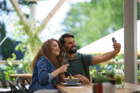Téléchargez les photos : Jeune couple à rendez-vous au restaurant, prenant selfie, assis sur la terrasse du restaurant. Petit ami et petite amie profitant du printemps, déjeuner ou brunch à l'extérieur, sièges extérieurs pour manger. - en image libre de droit