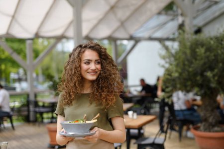 Foto de Retrato de una hermosa camarera sosteniendo el plato con comida, tazón con ensalada.Servidor femenino de pie en la terraza del restaurante en un delantal. - Imagen libre de derechos
