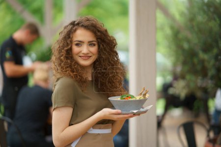 Foto de Retrato de una hermosa camarera sosteniendo el plato con comida, tazón con ensalada.Servidor femenino de pie en la terraza del restaurante en un delantal. - Imagen libre de derechos