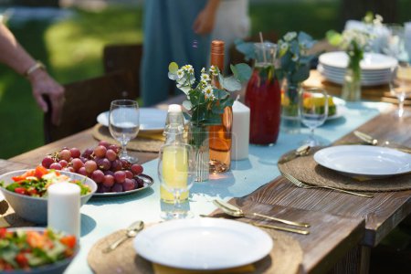Photo for Close up shot of a set table at summer garden party, grilled food. Table setting with glasses, lemonade, delicate floral and paper decoration, and bottles of summer wine. - Royalty Free Image