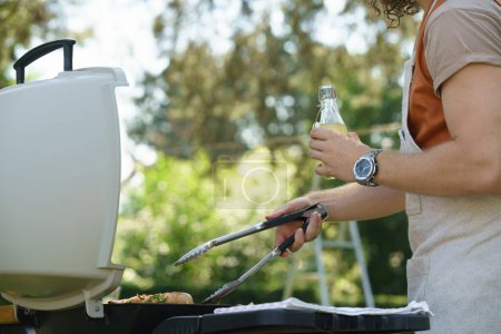 Man grilling on outdoor grill, holding tongs and drinkig lemonade. Outdoor grill or BBQ party in the garden.