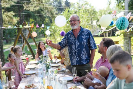 Abuelo tintineo con copa de champán. Hombre mayor de pie, haciendo un brindis de celebración en la fiesta al aire libre del jardín de verano.