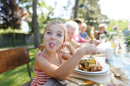 Foto de Retrato de una niña divertida sentada a la mesa comiendo comida a la parrilla al aire libre, sacando la lengua. Chica en la fiesta del jardín familiar. - Imagen libre de derechos