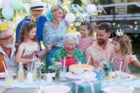 Photo for Garden birthday party for senior lady. Beautiful senior birthday woman receiving gift from granddaughter, family and friends. Birthday celebration with paper crowns. - Royalty Free Image