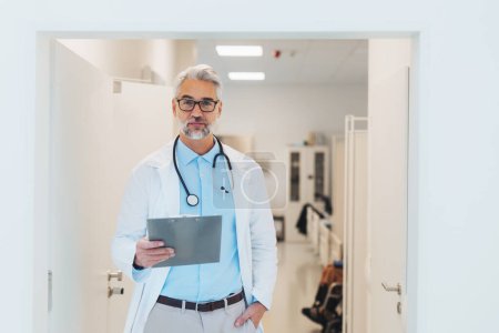 Portrait of mature doctor in hospital corridor. Handsome doctor with gray hair wearing white coat, stethoscope around neck standing in modern private clinic.