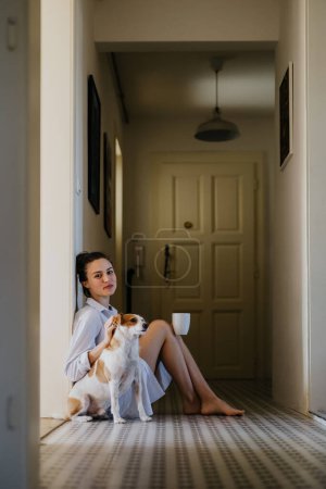 Foto de Mujer en pijama sentada en el suelo, jugando con su lindo perro, bebiendo una taza de café de la mañana. Relajación de fin de semana en casa. Estilo de vida Hygge. - Imagen libre de derechos