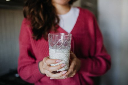 Close upf of woman in pink sweater holding glass with plant-based milk in the kitchen.