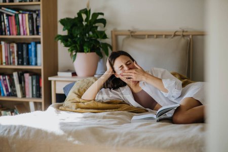 Foto de Hermosa mujer bostezando, leyendo libro en la cama, sensación acogedora. Libertad, actividad de fin de semana para mujer soltera. - Imagen libre de derechos