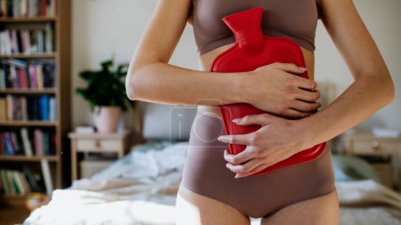 Mujer joven en casa sufriendo de dolor menstrual, con calambres. Mujer calentando la parte inferior del abdomen con una botella de agua caliente, endometriosis, y condiciones que causan dolor en la barriga. De cerca..