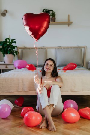 Photo for Beautiful woman sitting by bed in the middle of baloons, holding heart shaped balloon in hand. Concept of Valentines Day, love and romantic dating. - Royalty Free Image