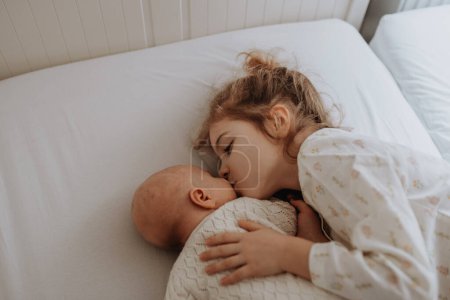 Porträt der großen Schwester beim Kuscheln mit Neugeborenem, kleinem Baby. Mädchen liegt mit ihrem neuen Geschwisterchen im Bett und küsst Babys Wange. Schwesternliebe, Freude für das neue Familienmitglied.