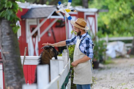 Foto de Retrato de granjero guapo acariciando cabra marrón en su granja. Hombre con sombrero trabajando en rancho familiar. - Imagen libre de derechos
