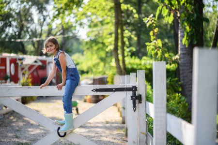 Téléchargez les photos : Belle jeune fille en bottes et salopettes en caoutchouc assis sur une clôture en bois blanc, aidant sur la ferme familiale pendant l'été. - en image libre de droit