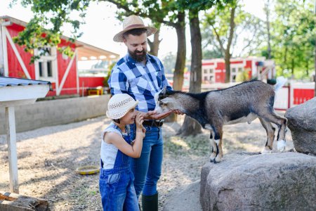 Photo for Portrait of father and young daughter standing in paddock, petting goat family farm. Concept of multigenerational farming. - Royalty Free Image