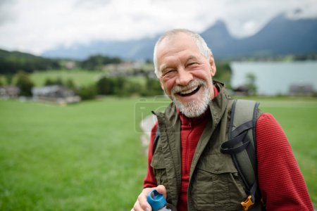 Téléchargez les photos : Portrait d'un homme âgé souriant marchant à l'extérieur avec des bâtons de trekking, allant sur un sentier de randonnée. Touristes seniors profitant de la vue. - en image libre de droit