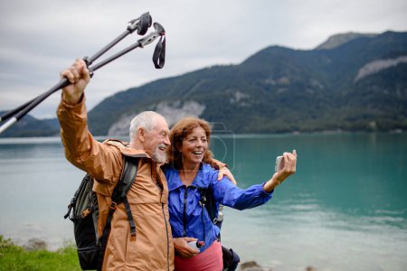 Foto de Retrato de una hermosa pareja de ancianos activos tomando selfie durante la caminata en las montañas de otoño. Turistas mayores abrazándose frente al lago. - Imagen libre de derechos