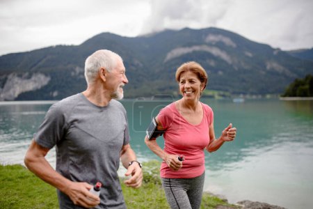 Photo for Senior couple running by the lake in nature. Elderly husband and wife spending active vacation in the mountains, enjoying physical activity and relaxation outdoors. - Royalty Free Image
