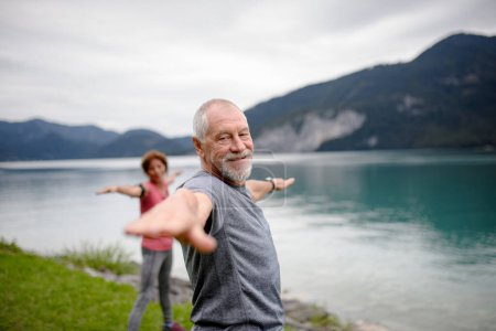 Téléchargez les photos : Couple de personnes âgées faisant du yoga en plein air, tai chi, pilates au bord du lac à l'automne. Le mari et la femme âgés passent des vacances actives dans les montagnes. Pensionnés étirant après l'entraînement extérieur. - en image libre de droit