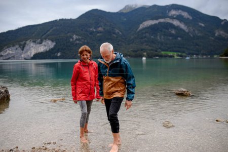 Active elderly couple hiking together in spring mountains, walking in cold fresh water in mountain lake. Senior tourists enjoying nature.