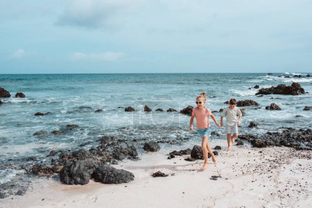 Photo for Siblings playing on beach, running, skipping rocks. Smilling girl and boy ion sandy beach with volcanic rocks of Canary islands. Concept of a family beach summer vacation with kids. - Royalty Free Image