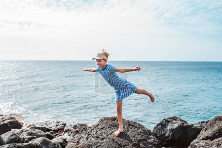 Young girl standing on one leg on rock. Blonde girl in dress and cap, visor enjoying vacation in Canary Islands. Concept of beach summer vacation with kids.