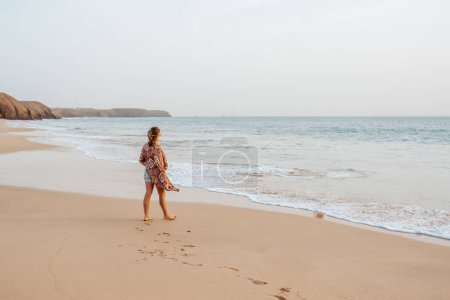 Téléchargez les photos : Vue arrière d'une belle femme mince debout sur la plage de sable. Plan complet du corps d'une femme pieds nus regardant la mer. Concept de vacances d'été à la plage. - en image libre de droit