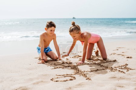 Photo for Siblings playing on beach, drawing tic-tac-toe in the sand, Smilling girl and boy in swimsuit on sandy beach of Canary islands. Concept of family beach summer vacation with kids. - Royalty Free Image