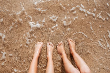 Téléchargez les photos : Gros plan des pieds sur la plage avec de l'eau de mer en dessous. Mousse de mer et jambes trempées d'eau chaude. - en image libre de droit