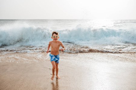 Photo for Young boy playing, running and splashing in strong sea waves. Smilling boy in swimsuit and swimming goggles. Concept of a beach summer vacation with kids. - Royalty Free Image