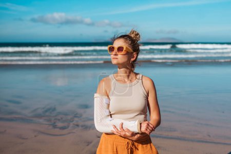 Photo for Beautiful woman with broken arm on beach. Arm cast, injured during family vacation in holiday resort. Concept of beach summer vacation. - Royalty Free Image