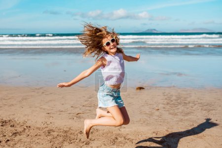 Photo for Jumping girl on the beach. Smilling blonde girl enjoying sandy beach, looking at crystalline sea in Canary Islands. Concept of beach summer vacation with kids. - Royalty Free Image
