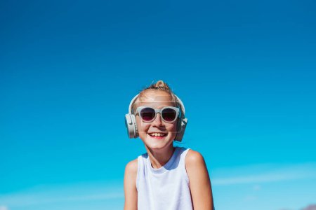 Photo for Girl with headphones on head standing on beach. Smilling blonde girl enjoying sandy beach, listening music via wireless headphones. Concept of beach summer vacation with kids. - Royalty Free Image