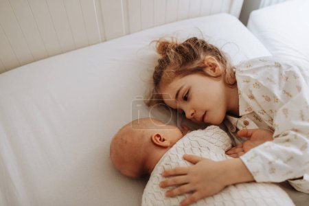 Foto de Retrato de la hermana mayor abrazando al recién nacido, bebé pequeño. Chica acostada con su nuevo hermano en la cama, ojos cerrados. Amor fraternal, alegría por el nuevo miembro de la familia. - Imagen libre de derechos
