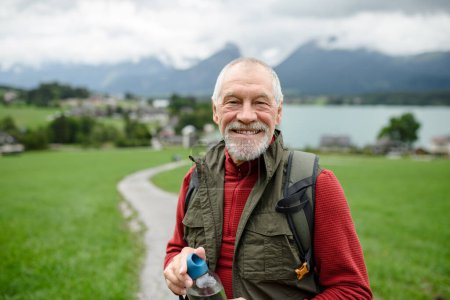 Téléchargez les photos : Portrait d'un homme âgé souriant marchant à l'extérieur avec des bâtons de trekking, allant sur un sentier de randonnée. Touristes seniors profitant de la vue. - en image libre de droit