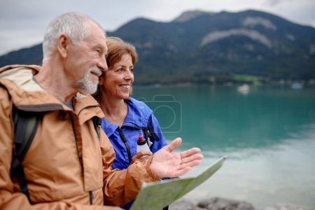 Foto de Retrato de una hermosa pareja activa de ancianos que caminan juntos en las montañas de primavera. Turistas mayores mirando el mapa frente a un lago. - Imagen libre de derechos