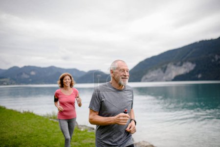 Photo for Senior couple running by the lake in nature. Elderly husband and wife spending active vacation in the mountains, enjoying physical activity and relaxation outdoors. - Royalty Free Image